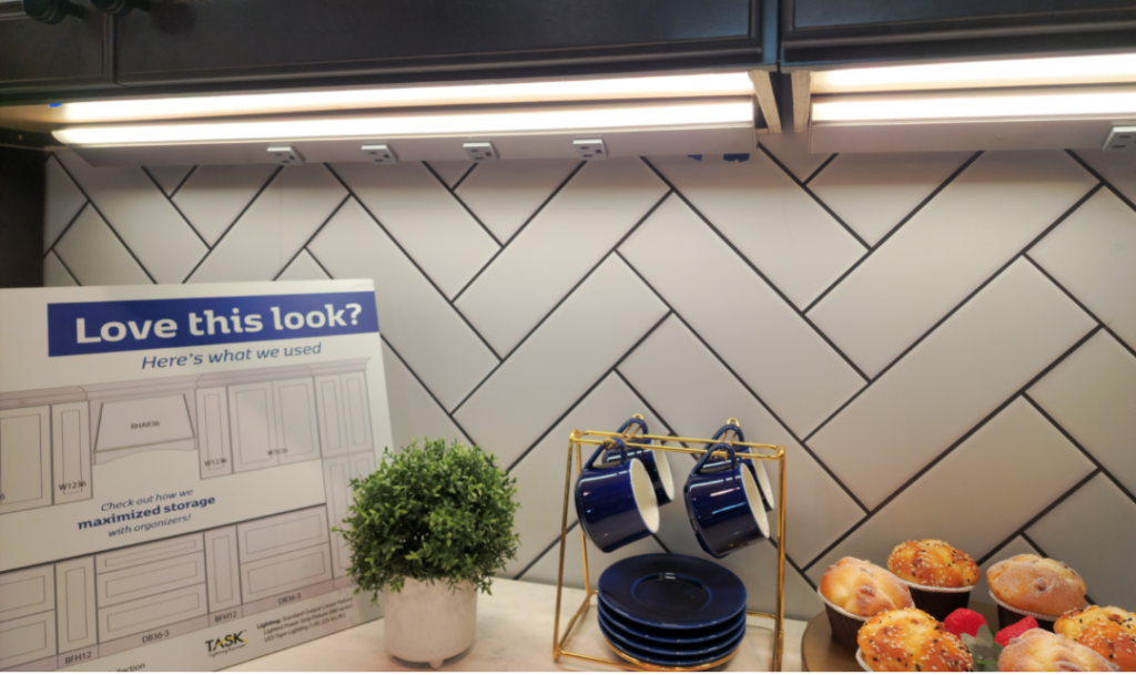 A kitchen display with two lighted powerstrips overhead, and a counter with a potted plant, blue mugs and plates, and a platter of muffins
