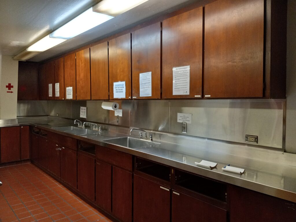 The back wall of the Community Kitchen remodel, before our team came in. Old red tile flooring, dark wood cabinets, outdated overhead lighting fixtures.